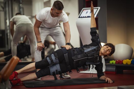 A smiling senior woman is doing plank exercises with personal trainer in the gym, dressed in a black suit with an EMS electronic simulator to stimulate their muscles.