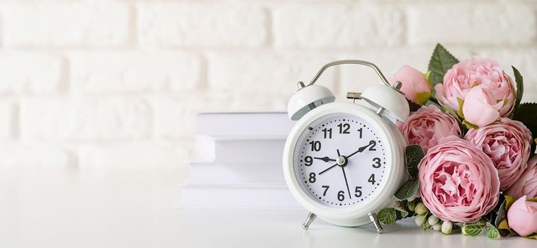 Time management, punctuality, business metaphors. Home interior. White vintage alarm clock on white brick wall background with stack of white books and flowers, copy space