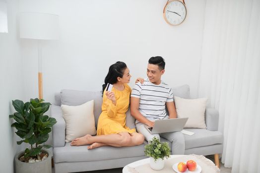 Choosing what to get. Beautiful young woman holding credit card and pointing laptop with smile while sitting together with her husband on the couch