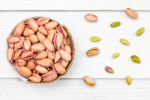 Overhead shot, roasted pistachios nuts in small bowl, some scattered on white boards desk.