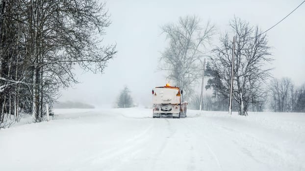 Snowplow highway maintenenace truck cleaning road completely white from snow in winter, dangerous driving conditions, view from car behind.