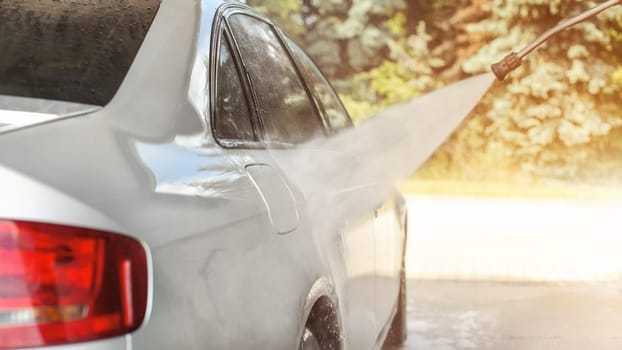 Silver car washed in self serve carwash, water spraying from the nozzle, sun lit trees in background.