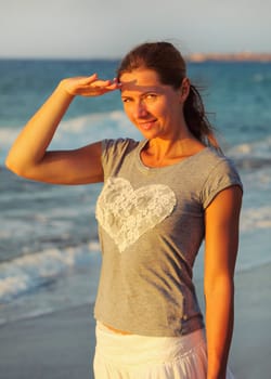 Young woman standing on the beach, shading her eyes from sun light during sunset, sea behind her
