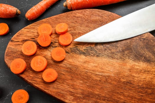 Wooden cutting board (space for text) with carrot sliced to small circles, blade of chef knife visible, view from above.