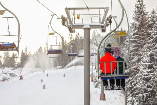 View from chairlift over ski piste, skier in bright red jacket seating in front, more blurred people skiing below, active snowguns background.