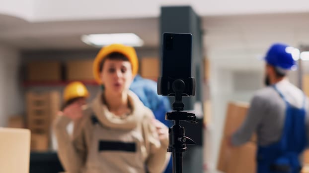 Caucasian woman recording video ad in warehouse, advertising merchandise on mobile phone online app. Young worker in overalls and hardhat filming social media video, industrial department.