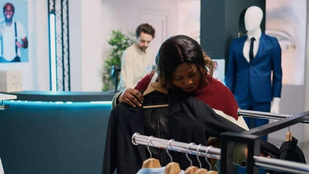 African american girl shopping for modern clothes on shop racks, looking to buy new fashion collection in mall boutique. Shopaholic woman checking fashionable merchandise, small business.