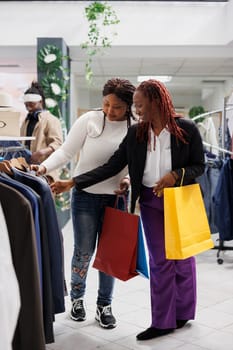 Two girlfriends carrying purchase bags and browsing jackets display rack in shopping center. Cheerful african american women with paper packages choosing blazer in fashion boutique
