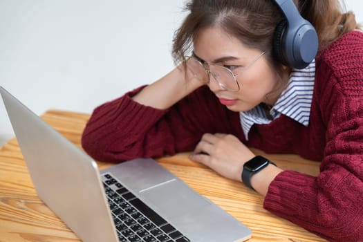 Asian college student at home wearing red shirt using laptop attending online university class listening with headphones.