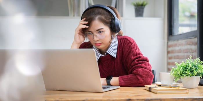 Asian college student at home wearing red shirt using laptop attending online university class listening with headphones.