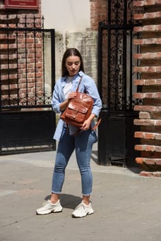 Beautiful young girl wearing blue jeans, shirt and white t-shirt posing with brown leather backpack. Street photo