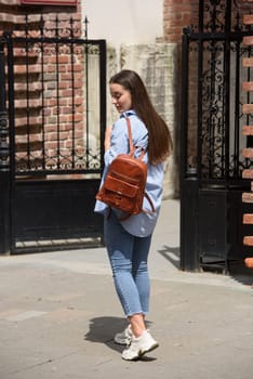 Beautiful young girl wearing blue jeans, shirt and white t-shirt posing with brown leather backpack. Street photo