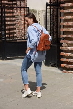 Beautiful young girl wearing blue jeans, shirt and white t-shirt posing with brown leather backpack. Street photo