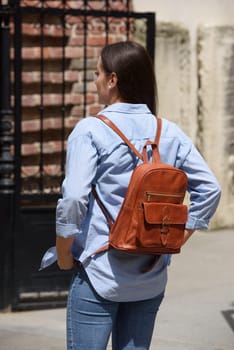 Beautiful young girl wearing blue jeans, shirt and white t-shirt posing with brown leather backpack. Street photo