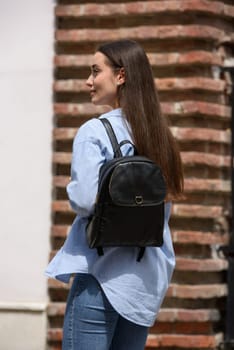 Beautiful young girl wearing blue jeans, shirt and white t-shirt posing with black leather backpack. Street photo