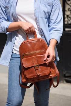 Beautiful young girl wearing blue jeans, shirt and white t-shirt posing with brown leather backpack. Street photo