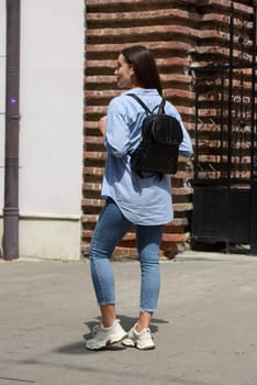 Beautiful young girl wearing blue jeans, shirt and white t-shirt posing with black leather backpack. Street photo