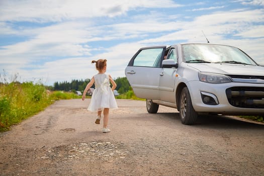 Portrait of little girl with Asian eyes on a road near car in a meadow or field with grass and flowers on sunny summer day. Rural, rustic journey