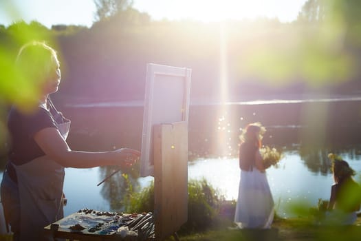 Adult female artist painting picture near water of river or lake in nature and girls in white sundress and flower wreath. Artist and models posing in holiday of Ivan Kupala in nature at sunset