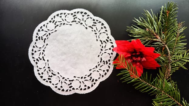 Black frame, white round napkin, green pine branch, red flower on background with reflection and sparkle of light. Abstract location for photo shoot. Cchristmas table decor. Concept of winter holiday