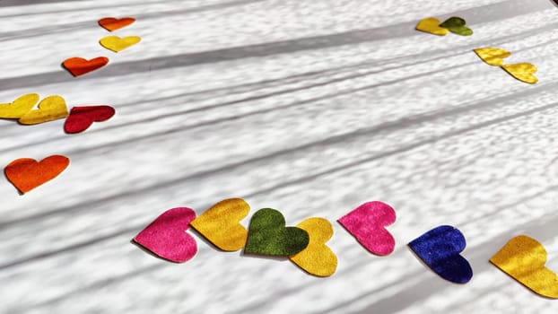 Multicolored hearts made of rough velvet paper on white surface with stripes of shadow and light from the sun. Abstract background, texture, copy space