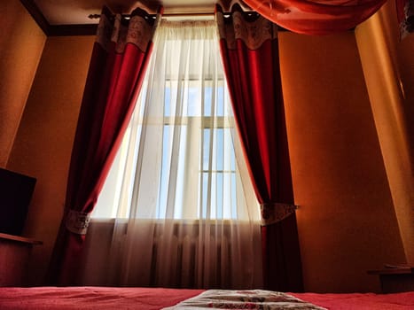 Beautiful red curtains on the window and transparent tulle fabric between them. View of daytime street. Background, texture, location, cozy room, and interior.