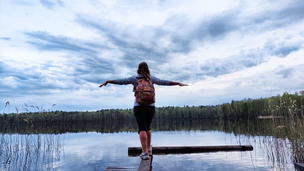 Girl or woman walks along old wooden pier, with aake in background during evening and landscape featuring water, forest and clouds in sky. Tourist in natural landscape. Concept of tourism and freedom