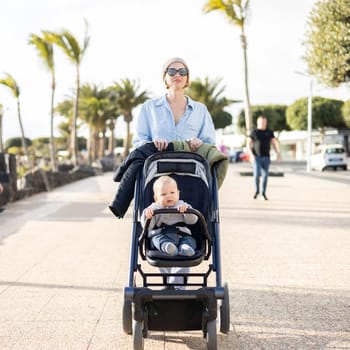 Fashinable mother walking and pushing her infant boy child in baby stroller along palm lined beach promenade of Puerto del Carmen, Lanzarote, Canary islands, Spain.