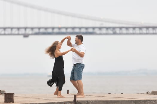 A romantic married couple dancing on the pier with a bridge on the background. Mid shot