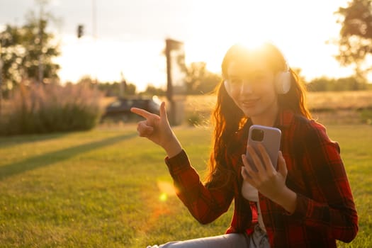 Woman sitting in the park and listening to music via headphone shows by finger to the left at sunlight