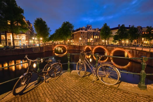Night view of Amsterdam cityscape with canal, bridge with bicycles and medieval houses in the evening twilight illuminated. Amsterdam, Netherlands