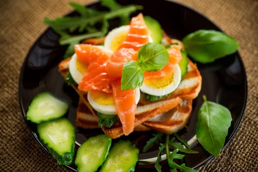 Cooked sandwich with lightly salted red fish, cucumbers and boiled eggs with herbs, in a plate.