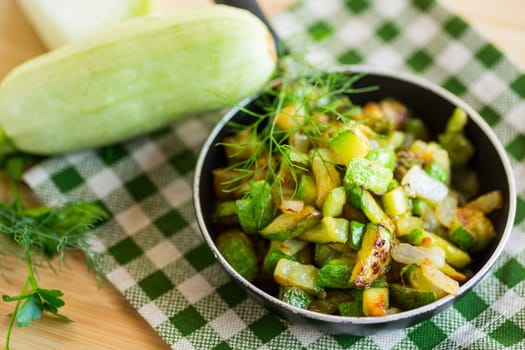 Sliced zucchini fried with onions and herbs in a frying pan on a wooden table.