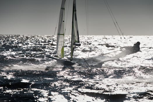 The view of sports sailboat is heeling at sunset, athletes tilt the boat, speed and spray, reflection of sun on water surface. High quality photo