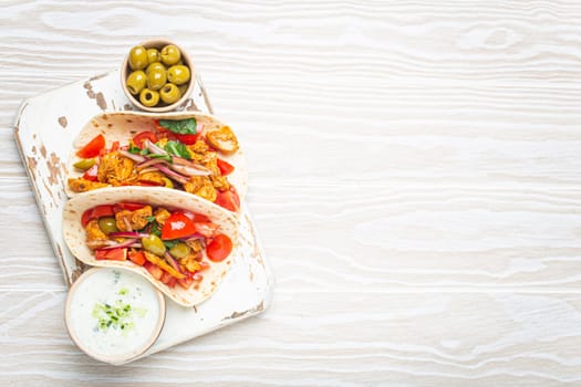 Traditional Greek Dish Gyros: Pita bread Wraps with vegetables, meat, herbs, olives on rustic wooden cutting board with Tzatziki sauce, olive oil top view on white wooden background, space for text.