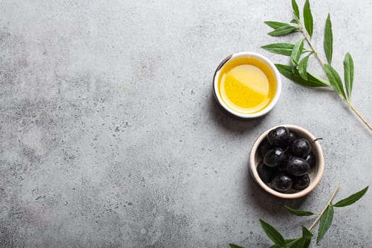 Black olives and extra virgin olive oil in little bowl with olive tree branches top view on gray concrete stone background, space for text