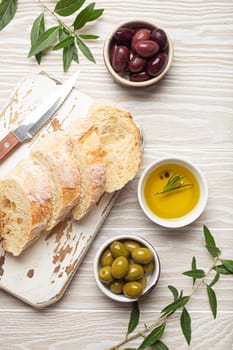 Sliced fresh ciabatta on cutting kitchen board, green and brown olives, olive oil with rosemary, olive tree branches, white wooden rustic background top view