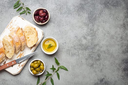 Sliced fresh ciabatta on cutting kitchen board, green and brown olives, olive oil with rosemary, olive tree branches on gray concrete stone rustic background from above copy space