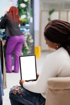 African american woman using tablet mock up while shopping in fashion boutique. Customer holding digital gadget with white empty screen in vertical mode while sitting in department store