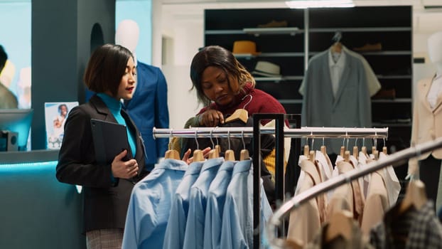 Group of people looking at fashion merchandise on racks, customer trying to buy new trendy clothes. Female shopper looking at formal wear in clothing store, talking to boutique employee.