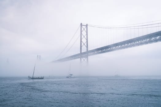 View of 25 de Abril Bridge famous tourist landmark of Lisbon connecting Lisboa and Almada in heavy fog mist wtih yacht boats passing under. Lisbon, Portugal