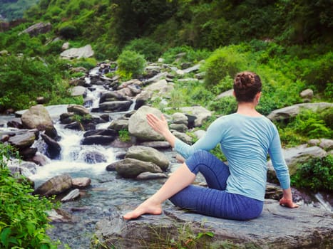 Yoga exercise outdoors - woman doing Ardha matsyendrasana asana - half spinal twist pose at tropical waterfall in Himalayas in India. Vintage retro effect filtered hipster style image.