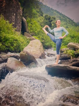 Yoga outdoors - woman doing Ashtanga Vinyasa Yoga balance asana Utthita Hasta Padangushthasana - Extended Hand-To-Big-Toe Pose position outdoors at waterfall. Vintage retro effect filtered image