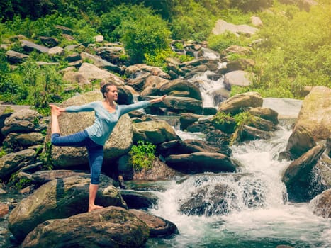 Yoga outdoors - woman doing yoga asana Natarajasana - Lord of the dance balance pose outdoors at waterfall in Himalayas. Vintage retro effect filtered hipster style image.