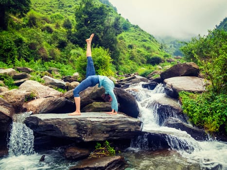 Yoga outdooors - woman doing yoga asana eka pada urdva dhanurasana Upward Bow Pose back benkd outdoors at waterfall in Himalayas. Vintage retro effect filtered hipster style image.