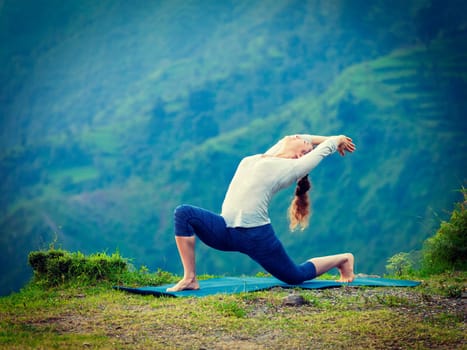 Yoga outdoors - sporty fit woman practices Hatha yoga asana Anjaneyasana - low crescent lunge pose posture outdoors in Himalayas mountains. Vintage retro effect filtered hipster style image.