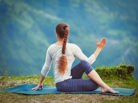 Hatha yoga outdoors - sporty fit woman doing yoga asana Parivrtta Marichyasana (or ardha matsyendrasana) - seated spinal twist outdoors in mountains. Vintage retro effect filtered hipster style image.