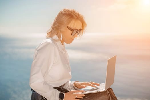 Business woman on nature in white shirt and black skirt. She works with an iPad in the open air with a beautiful view of the sea. The concept of remote work