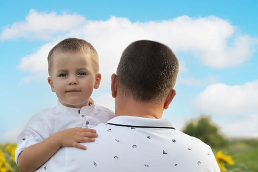 Father's day concept. A little boy in his father's arms, in white clothes, gently hugs and cuddles, against the blue sky, in a field with yellow sunflowers. A symbol of peace. Symbol of Ukraine. Close-up. copy space.