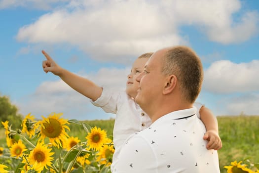 Father's day concept. Family. A little boy in his father's arms against a blue sky in a field with yellow sunflowers. The son stretched out his hand to the sky and points to his father in the sun. A symbol of peace. Symbol of Ukraine. Close-up. copy space.